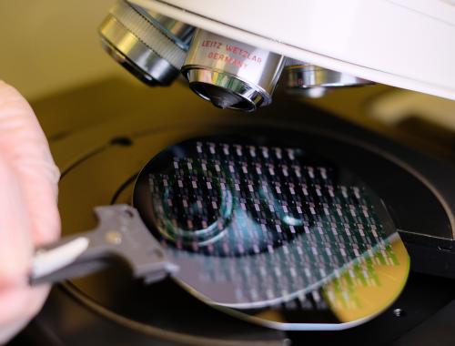 Una mano sujeta una oblea procesada con pinzas debajo de un microscopio en la Sala Blanca