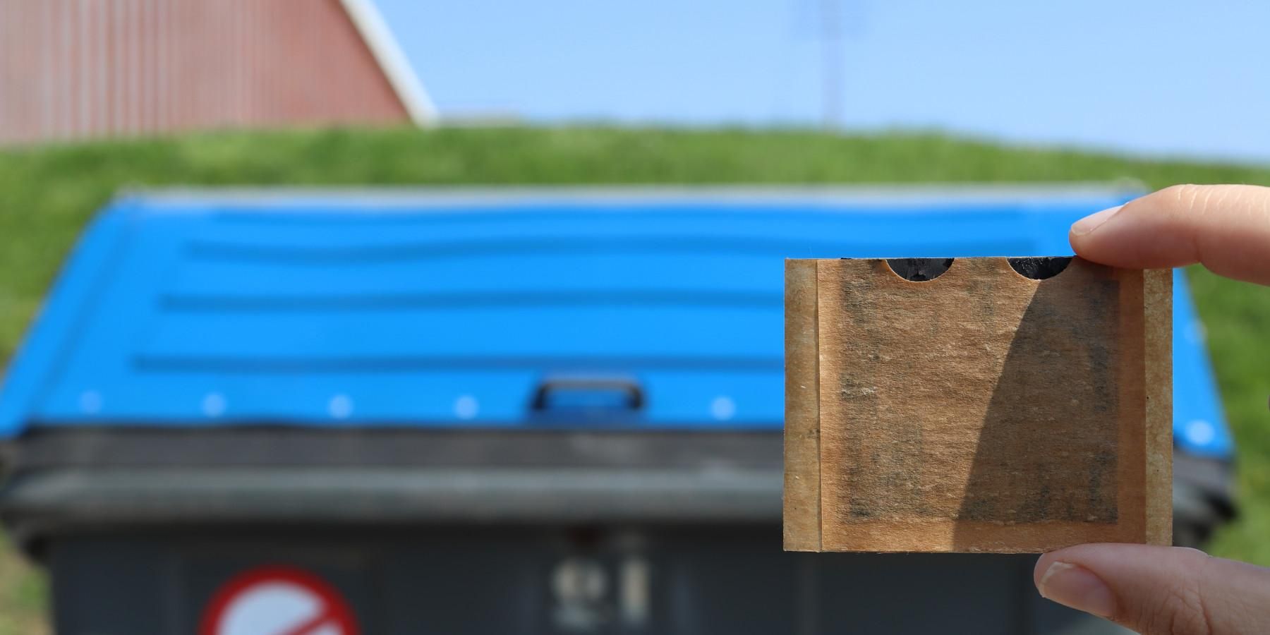 Una mano sostiene la batería adhesiva delante de un contenedor azul para reciclar papel y cartón. Crédito: Carles Tortosa y Marina Navarro.