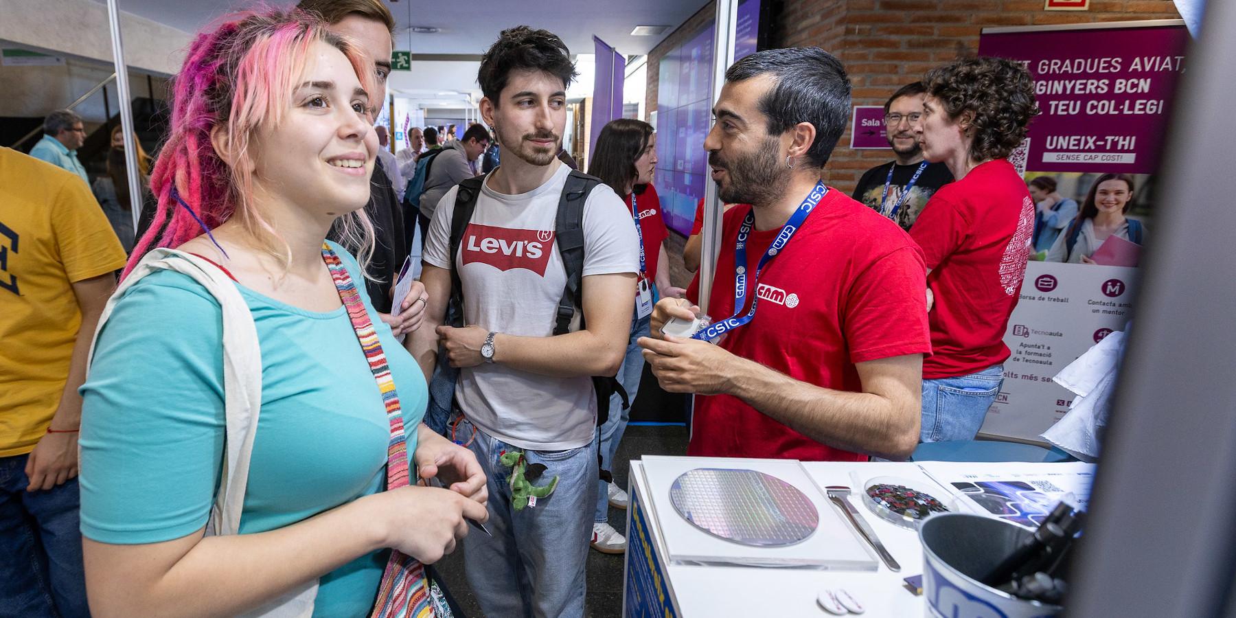 Chica en el stand del IMB-CNM preguntando con interés sobre el centro junto a dos amigos con personal del IMB-CNM atendiendo