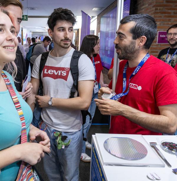Chica en el stand del IMB-CNM preguntando con interés sobre el centro junto a dos amigos con personal del IMB-CNM atendiendo